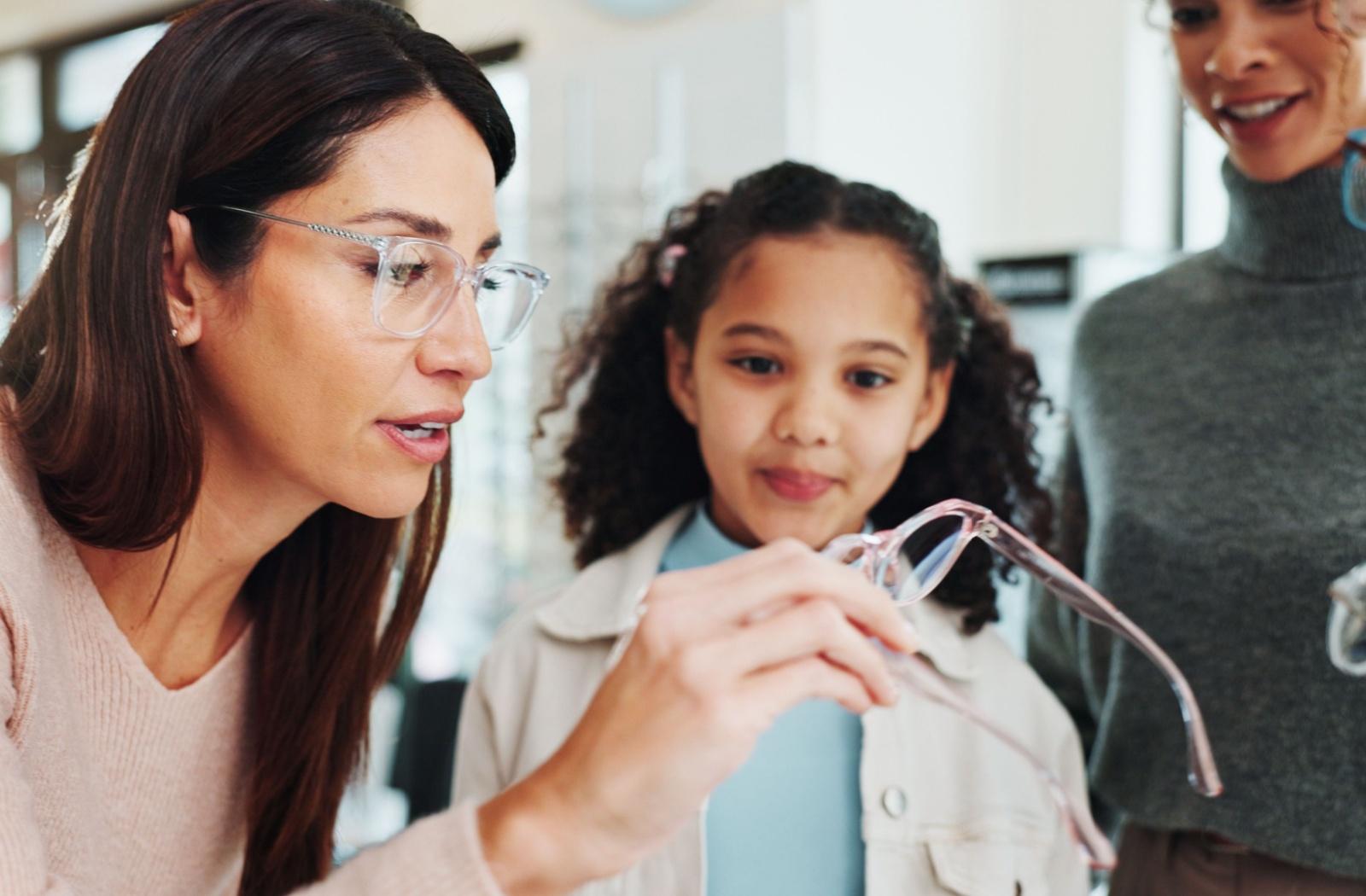 A child looking at a pair of pink glasses with an optician helping